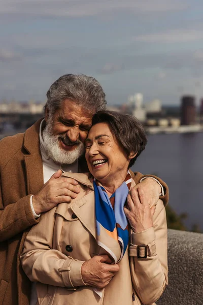 Uomo gioioso sorridente mentre abbraccia gioiosa moglie anziana in trench — Foto stock