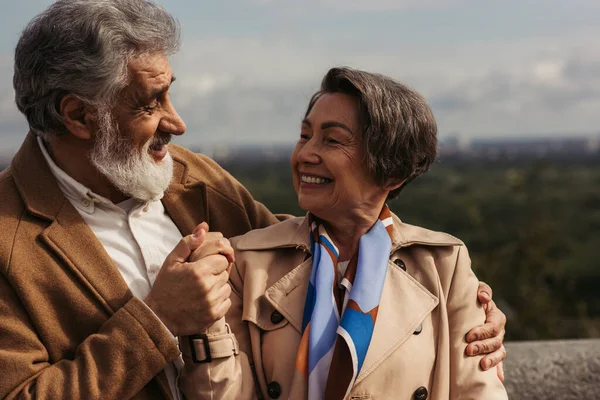 Homme barbu souriant tout en étreignant et en tenant la main avec la femme aînée en trench coat — Photo de stock