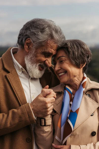 Uomo allegro sorridente mentre abbraccia e si tiene per mano con moglie anziana in trench — Foto stock