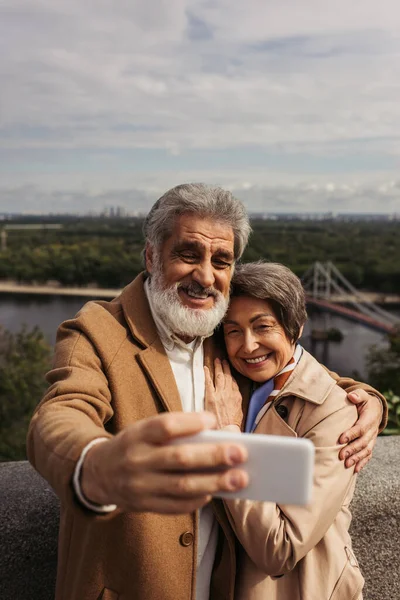 Happy senior man taking selfie with cheerful wife in trench coat near river — Stock Photo