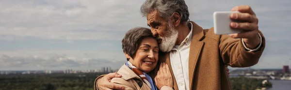 Homme âgé barbu embrassant la tête de femme gaie et prenant selfie, bannière — Photo de stock