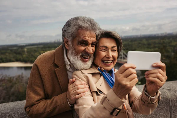 Heureuse femme âgée en trench coat et prendre selfie avec mari barbu — Photo de stock