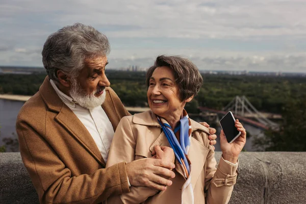 Mujer mayor feliz en gabardina beige mirando al marido barbudo y sosteniendo el teléfono inteligente con pantalla en blanco - foto de stock