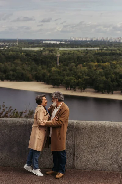 Piena lunghezza di allegra coppia anziana in cappotti autunnali beige che si abbracciano sul ponte vicino al fiume — Foto stock