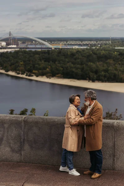 Glückliches Senioren-Paar in beigen Herbstmänteln umarmt sich auf Brücke am Fluss — Stockfoto