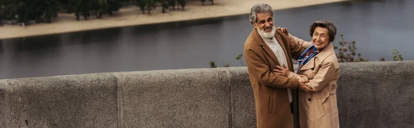 Cheerful senior couple in beige autumnal coats hugging on bridge near river, banner — Stock Photo