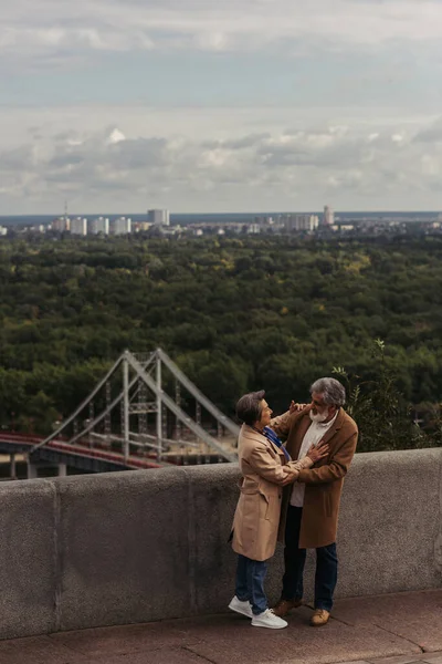 Cheerful senior couple in beige autumnal coats hugging on bridge near river — Stock Photo