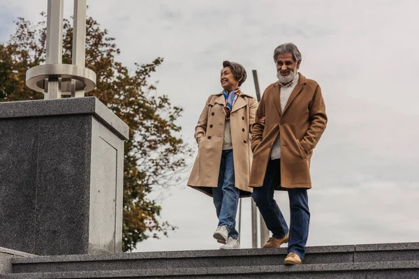 Full length of stylish senior man in coat walking on stairs with hands in pockets near happy wife — Stock Photo