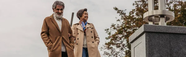 Hombre mayor con estilo en abrigo caminando con las manos en los bolsillos cerca de esposa feliz, bandera - foto de stock