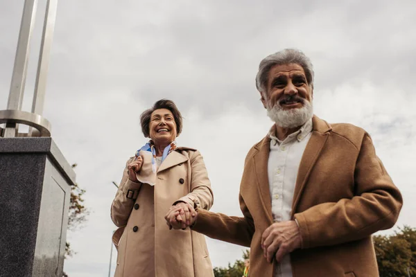Heureux senior homme dans manteau marche et tenant la main de femme heureuse contre ciel nuageux — Photo de stock