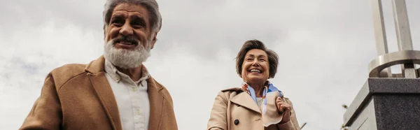 Zufriedener älterer Herr im Mantel lächelnd mit glücklicher Frau vor bewölktem Himmel, Banner — Stockfoto