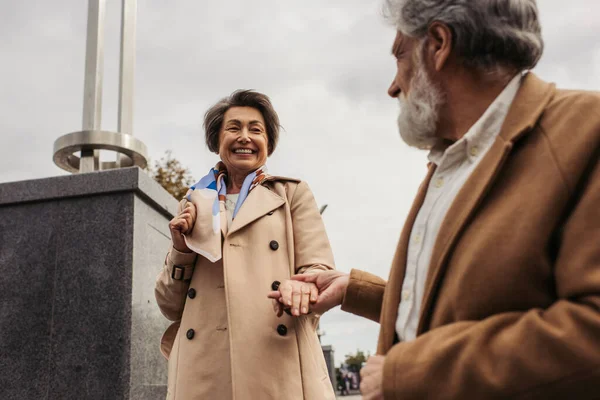 Homme âgé flou en manteau tenant la main de la femme heureuse contre le ciel nuageux — Photo de stock