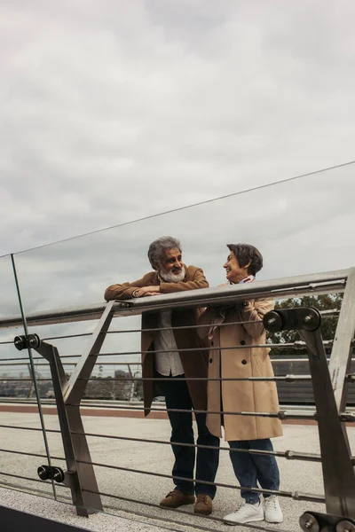 Cheerful senior man in coat looking at pleased wife while leaning on guard rail of bridge — Stock Photo