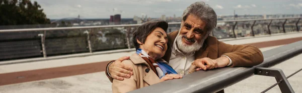Homme âgé heureux en manteau étreignant femme joyeuse tout en se tenant près du rail de garde du pont, bannière — Photo de stock