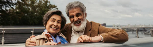 Uomo anziano barbuto in cappotto abbracciando moglie allegra e in piedi vicino ponte guardia rail, banner — Foto stock