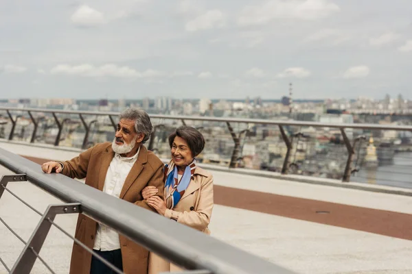 Happy senior man in coat and cheerful woman standing near bridge guard rail — Stock Photo