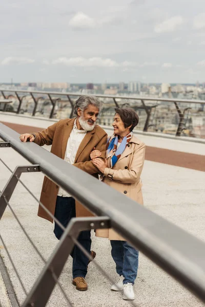 Feliz anciano mujer abrazando barbudo marido cerca puente guardia rail - foto de stock
