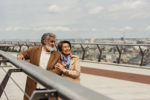 Glückliche Seniorin umarmt bärtigen und fröhlichen Ehemann in der Nähe der Brückenleitplanke — Stockfoto