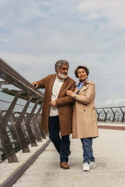 Full length of happy senior woman hugging bearded and cheerful husband near bridge guard rail — Stock Photo