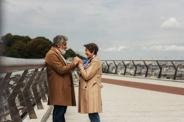 Vue latérale de la femme âgée heureuse tenant la main avec mari barbu et joyeux près du pont garde-corps — Photo de stock