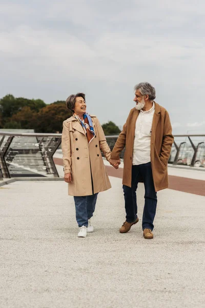 Longitud completa de alegre pareja de ancianos en abrigos tomados de la mano y caminando en el puente cerca de la barandilla de guardia - foto de stock