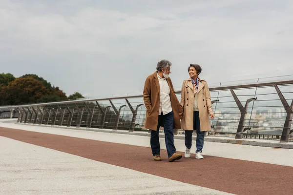 Longitud completa de la complacida pareja de ancianos en abrigos beige tomados de la mano y caminando en el puente cerca de carril de guardia - foto de stock