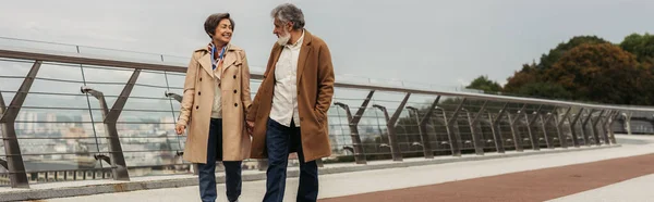 Gioiosa coppia anziana in cappotti beige che si tiene per mano e cammina sul ponte vicino al guard rail, striscione — Foto stock