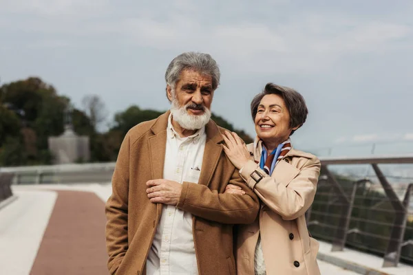 Cheerful senior couple in beige autumnal coats smiling while walking on bridge — Stock Photo