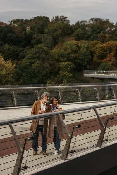 Eine glückliche Seniorin und ein bärtiger Mann stehen in voller Länge in der Nähe der Brückenleitplanke — Stockfoto