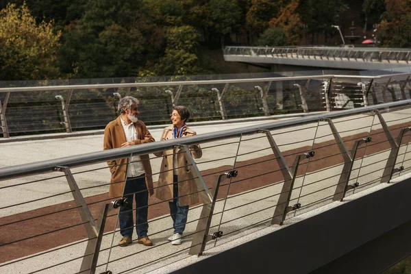 Volle Länge der glücklichen Seniorin und des bärtigen lächelnden Mannes, der in der Nähe der Brückenleitplanke steht — Stockfoto