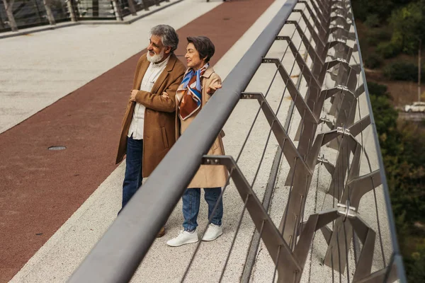 Longitud completa de la sonriente mujer mayor abrazando marido barbudo en el puente cerca de carril de guardia - foto de stock