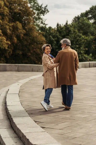 Piena lunghezza di donna anziana sorridente abbracciando il marito barbuto in cappotto e camminando fuori — Foto stock