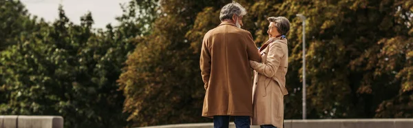Alegre anciano mujer abrazando barbudo marido en abrigo y de pie en parque, pancarta - foto de stock