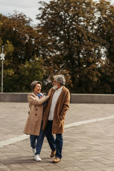 Fröhliche Seniorin umarmt bärtigen Mann im Mantel und steht im Stadtpark — Stockfoto