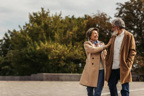 Joyful senior woman hugging bearded husband in beige coat and standing in park — Stock Photo