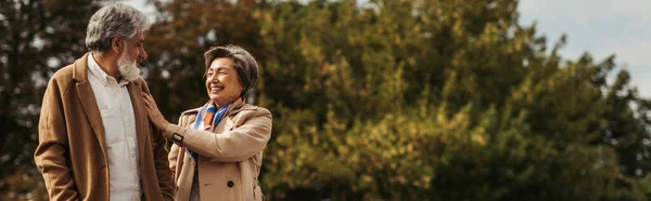 Joyeuse femme âgée étreignant mari barbu en manteau et debout dans le parc, bannière — Photo de stock