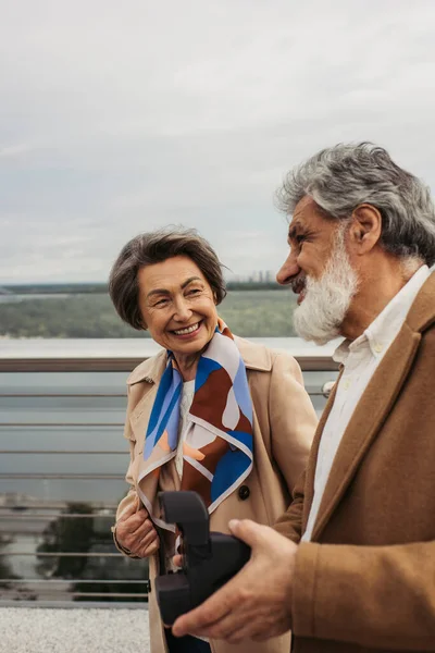 Щасливий старший чоловік тримає старовинну камеру біля цікавої дружини, посміхаючись на мосту — стокове фото