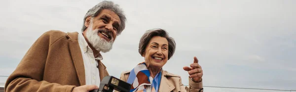 Бородатый и старший мужчина держит винтажную камеру рядом с женой улыбаясь, указывая пальцем, баннером — стоковое фото