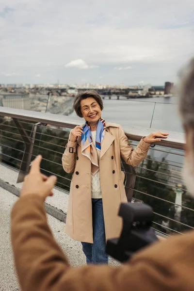 Homme flou tenant caméra vintage et geste près de femme âgée heureuse posant près du pont garde-corps — Photo de stock