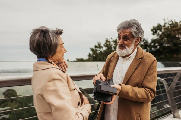 Anziano barbuto uomo tenendo fotocamera vintage e parlando con moglie in piedi vicino ponte guardia rail — Foto stock