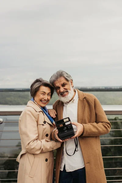 Старший бородатый мужчина держит винтажную камеру возле счастливой жены, стоя рядом с ограждением моста — стоковое фото