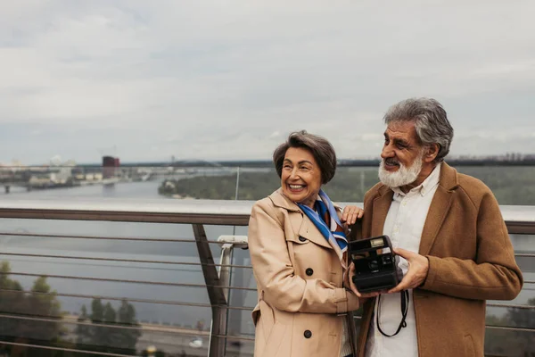 Mujer mayor feliz en gabardina abrazando marido con cámara vintage de pie fuera - foto de stock