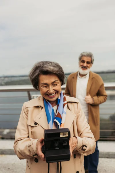 Happy senior woman holding vintage camera near blurred husband on background — Stock Photo