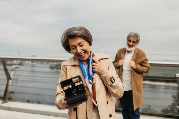 Mulher sênior alegre segurando câmera vintage perto do marido borrado no fundo — Fotografia de Stock