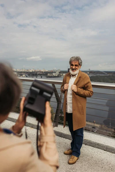 Blurred woman holding vintage camera while taking photo of bearded senior husband on bridge near river — Stock Photo