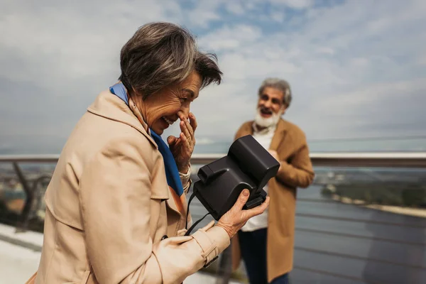 Positivo anziano donna holding vintage fotocamera e ridere vicino offuscata marito su sfondo — Foto stock