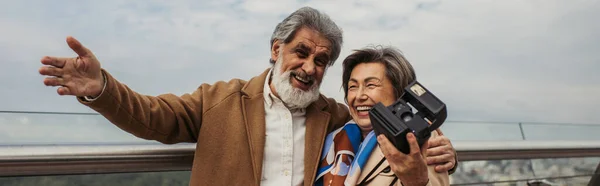 Alegre anciana sosteniendo la cámara vintage mientras su marido sonríe y gesticulando fuera, pancarta - foto de stock