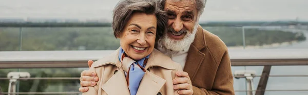 Alegre hombre con barba abrazando esposa mayor en gabardina exterior, pancarta - foto de stock