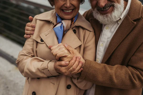 Vista ritagliata dell'uomo allegro con la barba che tiene la mano della moglie anziana in trench all'esterno — Foto stock