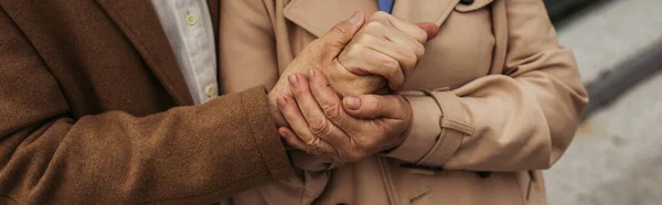 Обрезанный вид пожилого мужчины, держащего за руку жену в плаще снаружи, баннер — стоковое фото